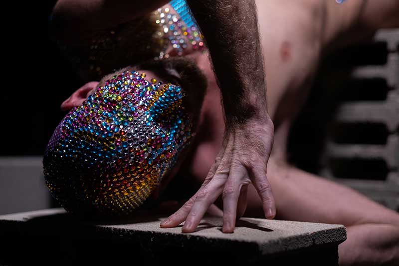 A imagem mostra um homem deitado em um bloco de concreto, com uma máscara de strass na cabeça. O homem é de pele branca, com cabelos curtos e barba. Ele está nu. A máscara é preta e branca, com strass brilhantes. O homem está olhando para a câmera com um sorriso no rosto.A imagem foi tirada em uma sala escura, com apenas uma luz direcional iluminando o homem. O fundo da imagem é preto.